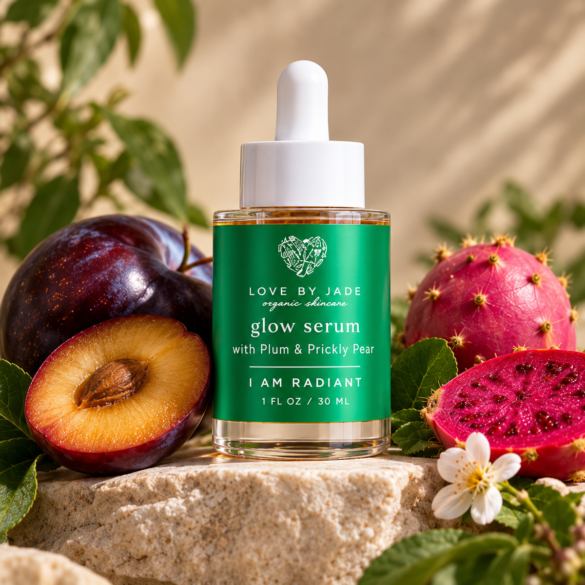 A bottle of Love By Jade Glow Serum is displayed on a stone pedestal surrounded by fresh ingredients, including a halved plum, a whole dragon fruit, and a white flower, with a soft-focus background of green leaves and a beige wall.